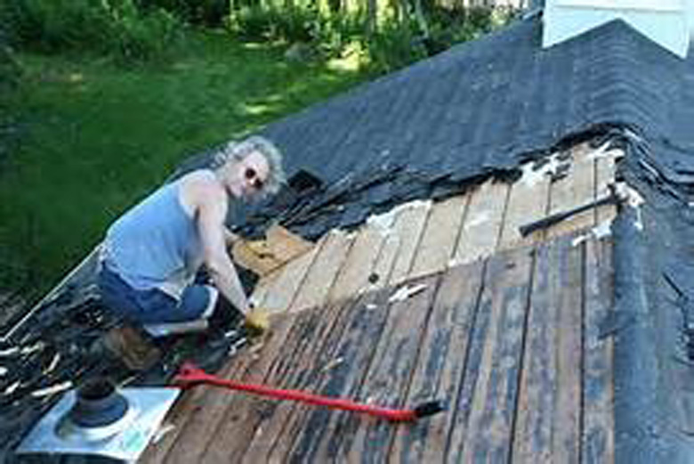 woman on in a blue shirt on top of a roof, removing roof shingles with a roof shovel